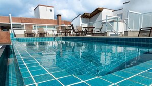 A swimming pool with blue tiles, surrounded by lounge chairs and tables, with a building in the background. - Apto a 280m da Praia do Itagu c Piscina e Garagem (Ubatuba)