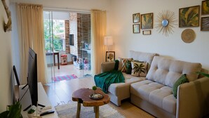 A living room with a sofa, a coffee table, a TV, and a balcony with a glass door. - Apto a 280m da Praia do Itagu c Piscina e Garagem (Ubatuba)