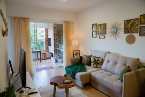 Uma sala de estar com sofá, mesa de centro, televisão e varanda com porta de vidro.