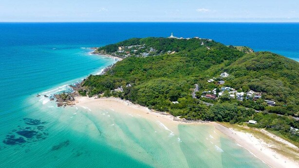 An aerial view of a coastal area with a beach, a lighthouse, and a hill with vegetation. - Byron Social on Jonson (Byron)