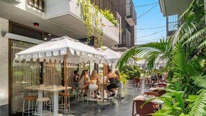 An outdoor cafe with tables and chairs, surrounded by greenery and a modern building. - Byron Social on Jonson (Byron)