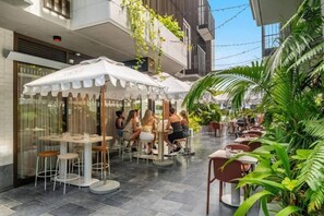 Un café en plein air, avec des tables et des chaises, entouré de verdure et bordé d’un bâtiment moderne.