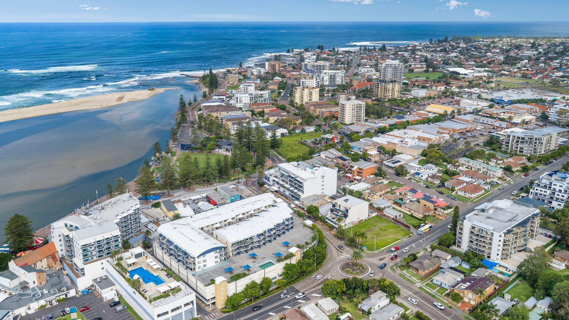 Eine Luftaufnahme einer Küstenstadt mit Strand, Wohngebäuden und einem großen Hotel mit Swimmingpool.