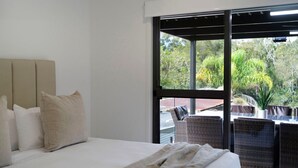 A bedroom with a bed, pillows, a window, and a view of outdoor furniture and greenery.