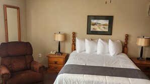 A hotel room with a bed, a chair, a ceiling fan, a nightstand, a lamp, a framed picture on the wall, and a carpeted floor.