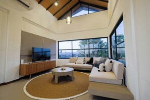 Ein modernes Wohnzimmer mit großem Fernseher, einer Couch, einem Couchtisch und Blick durch große Fenster.
