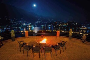 A terrace with a fire pit, chairs, and a view of a cityscape at night. - RnR Premium Stay (Bhimtal)