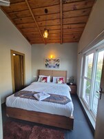 A bedroom with a wooden ceiling, a bed with white bedding, a nightstand with a lamp, and a window with a view.