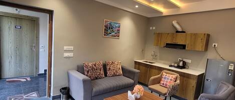 A modern living room with a sofa, armchair, coffee table, and a kitchen area with a refrigerator and sink.