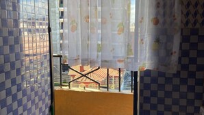Un balcon donnant sur des immeubles, un sol carrelé et une fenêtre ornée d’un rideau à motifs de fruits.