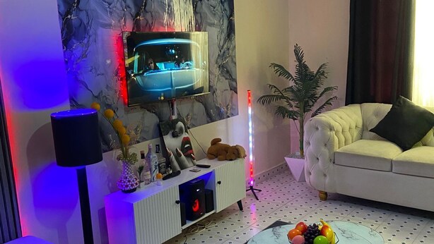 Un salon moderne avec un téléviseur à écran plat, un canapé blanc, une table basse ornée de fruits et une plante décorative.