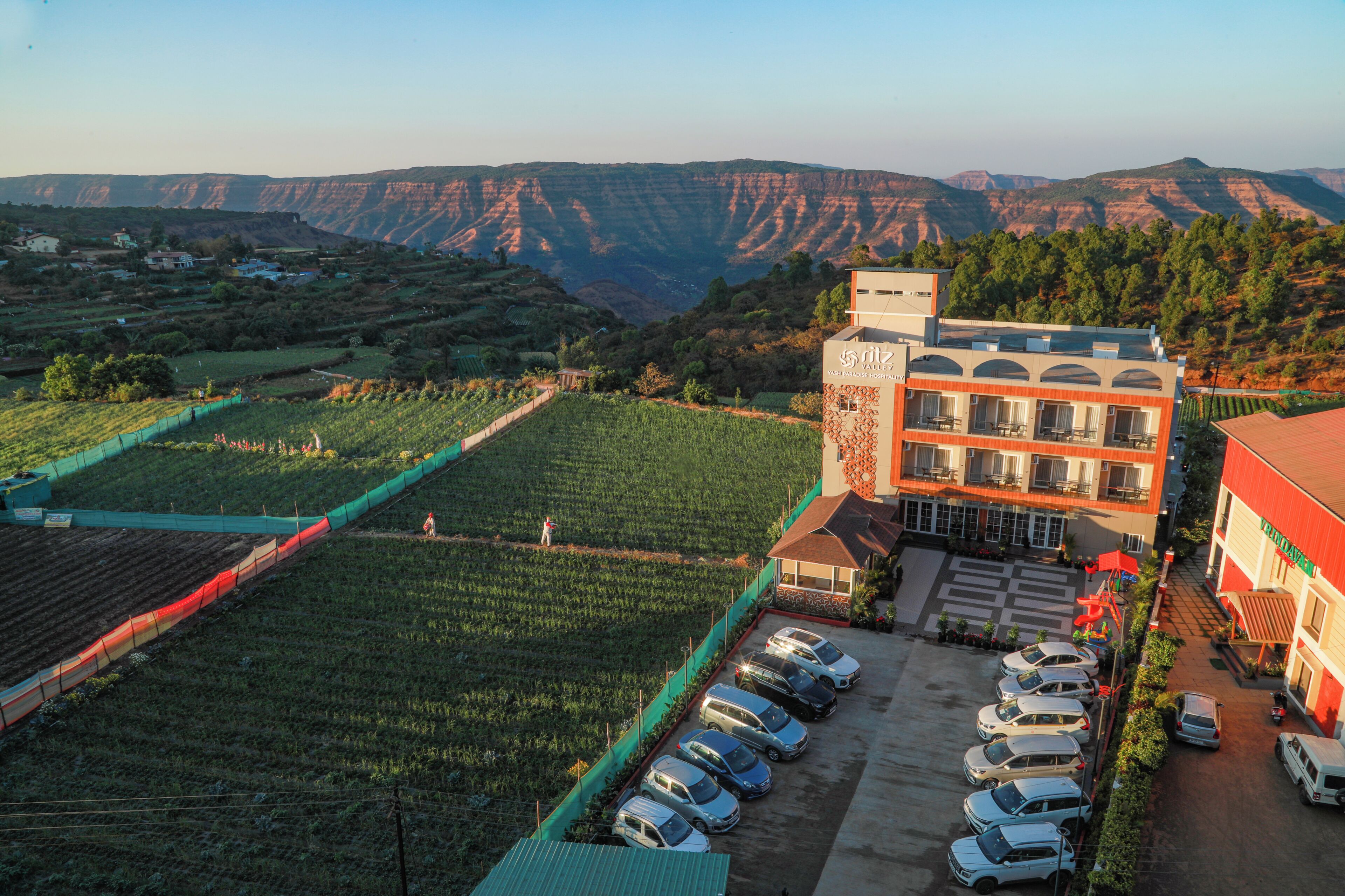 A hotel with a parking lot, a building with a red roof, and a view of a valley with terraced fields.