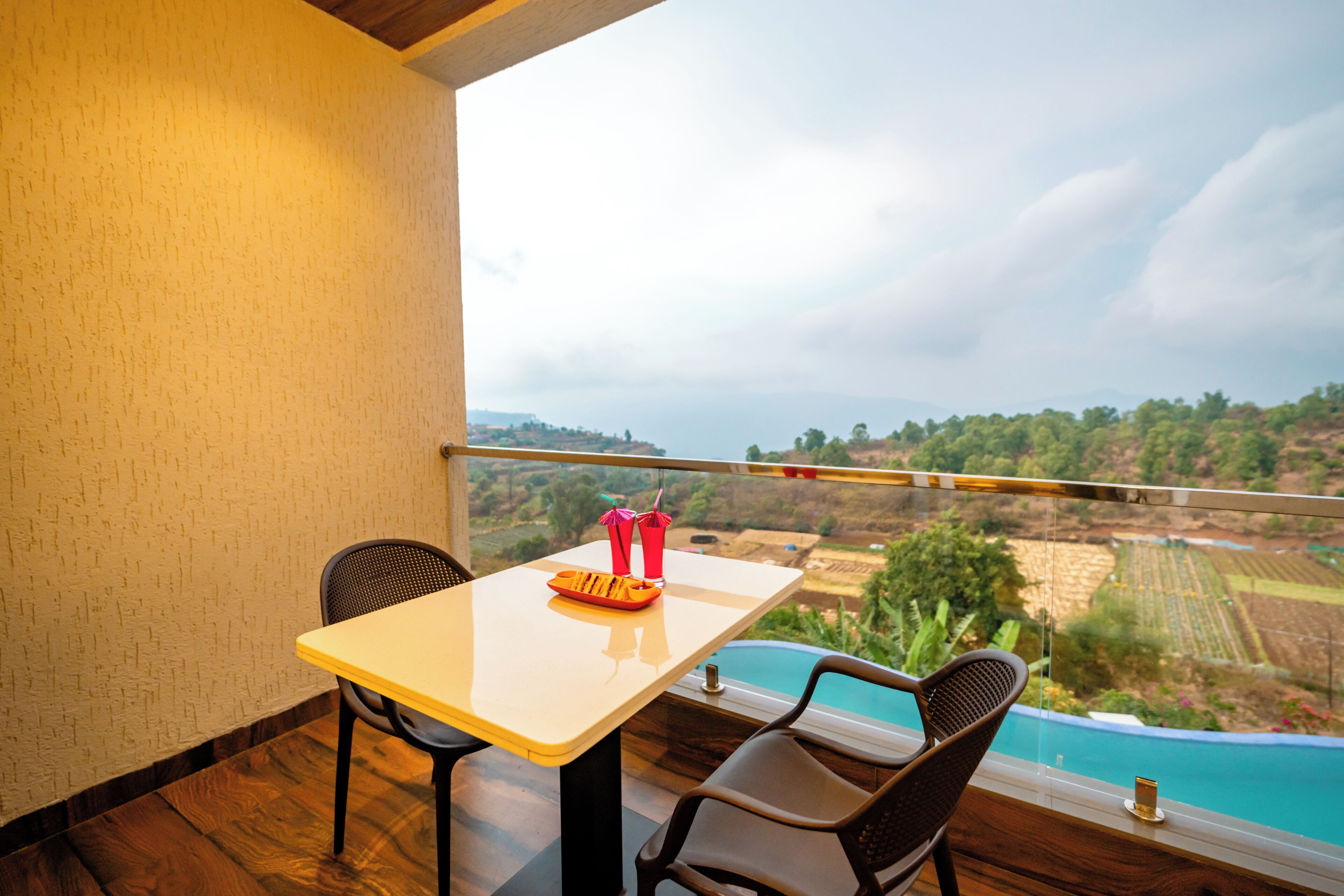 A balcony with a table and chairs, a view of a swimming pool, and a distant landscape.