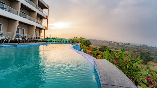 A swimming pool with a curved edge, a multi-story building with balconies, and a view of a landscape in the background. - Ritz Valley Resort (Mahabaleshwar)