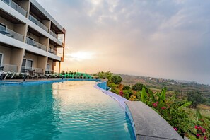 A swimming pool with a curved edge, a multi-story building with balconies, and a view of a landscape in the background. - Ritz Valley Resort (Mahabaleshwar)