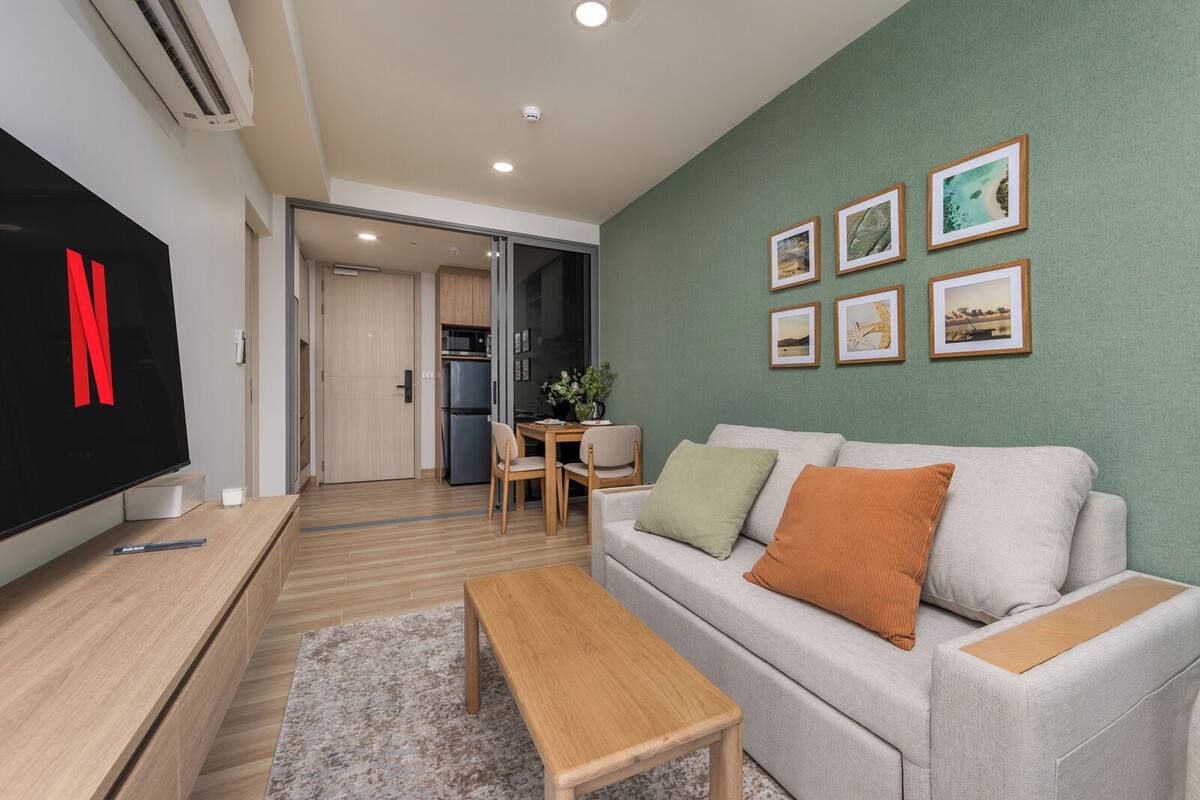 Una sala de estar moderna con un televisor de pantalla plana, un sofá gris con cojines, una mesa de centro de madera, un comedor con mesa y sillas, y una cocina al fondo.