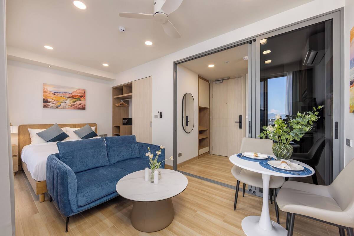 Una habitación de hotel moderna con una cama, un sofá azul, una mesa de centro redonda, una pequeña mesa de comedor con sillas y un ventanal corredero que da a un balcón.