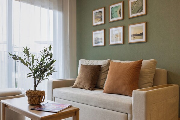 A living room with a beige sofa, two pillows, a small table with a plant and magazine, and framed pictures on the wall. - Skypark Celeste Laguna by TropicLook (Choeng Thale)