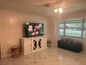 A living room with a flat-screen TV, a white cabinet with glass doors, a ceiling fan, a window with blinds, and a dark-colored sofa with colorful cushions. - Casa las Brisas, with an outdoor hot tub and a lovely place to relax  (Lehigh Acres)
