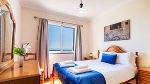 Habitación de hotel con cama, escritorio, silla, ventana con cortinas, un cuadro en la pared y un jarrón con flores.