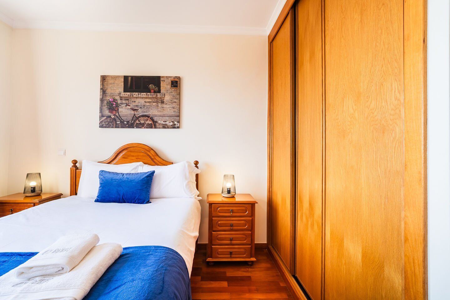 A hotel room with a wooden bed, a nightstand with a lamp, a wooden wardrobe, and a painting on the wall.