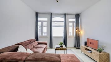 Una sala de estar moderna con un sofá seccional marrón, un televisor sobre un mueble de madera y ventanas amplias con cortinas.