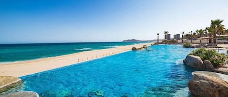 A clear blue pool adjacent to a sandy beach with a view of the ocean and distant mountains.