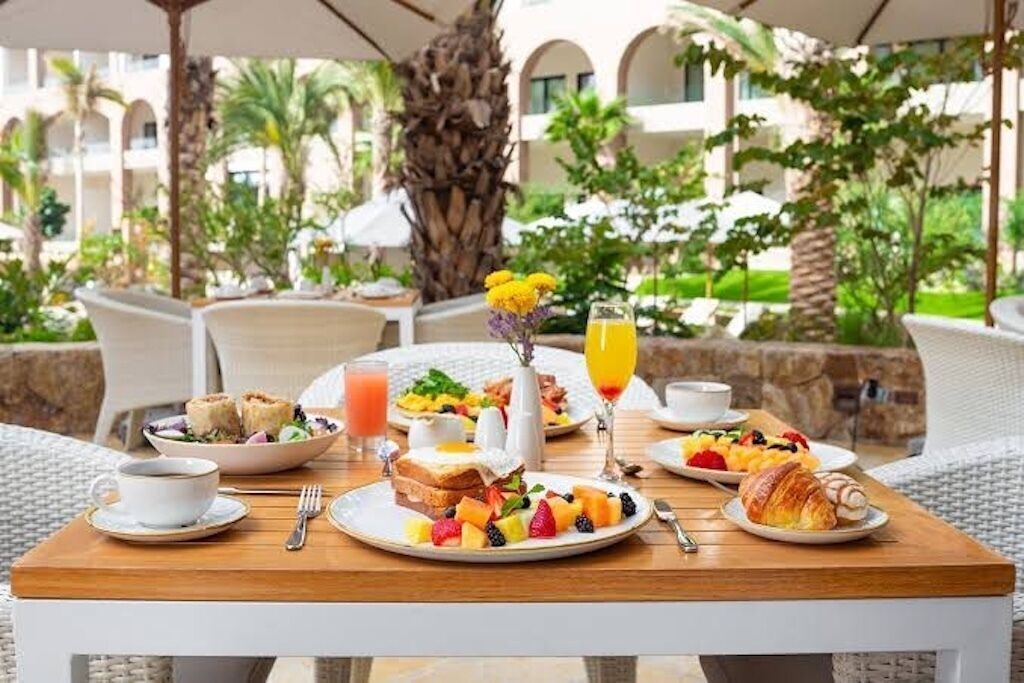 An outdoor dining area with a wooden table, plates of breakfast food, and a tropical garden view.