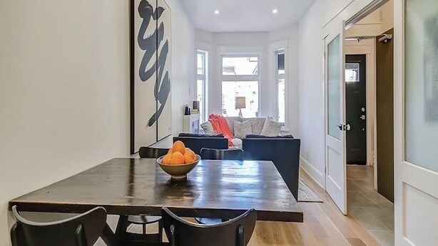 A modern dining area with a wooden table, chairs, a bowl of fruit, and a large abstract painting on the wall.
