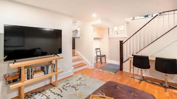 Uma sala de estar com televisão de tela plana, estantes de madeira, um banquinho de couro marrom, um tapete estampado e uma escada que leva a um andar superior.