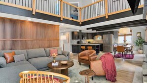 Een ruime woonkamer met een grijze bank, een wand bekleed met hout, een eethoek met tafel en stoelen, en een keuken met moderne apparatuur.