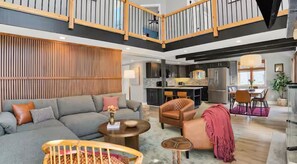 Ein großzügiger Wohnbereich mit grauem Sofa, einer Wandpaneele aus Holz, einer Essecke mit Tisch und Stühlen sowie einer Küche mit modernen Geräten.