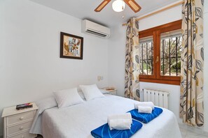 Ein ordentlich eingerichtetes Schlafzimmer mit Bett, Nachttischen, einer Klimaanlage, einem Fenster mit Vorhängen und einem Heizkörper.