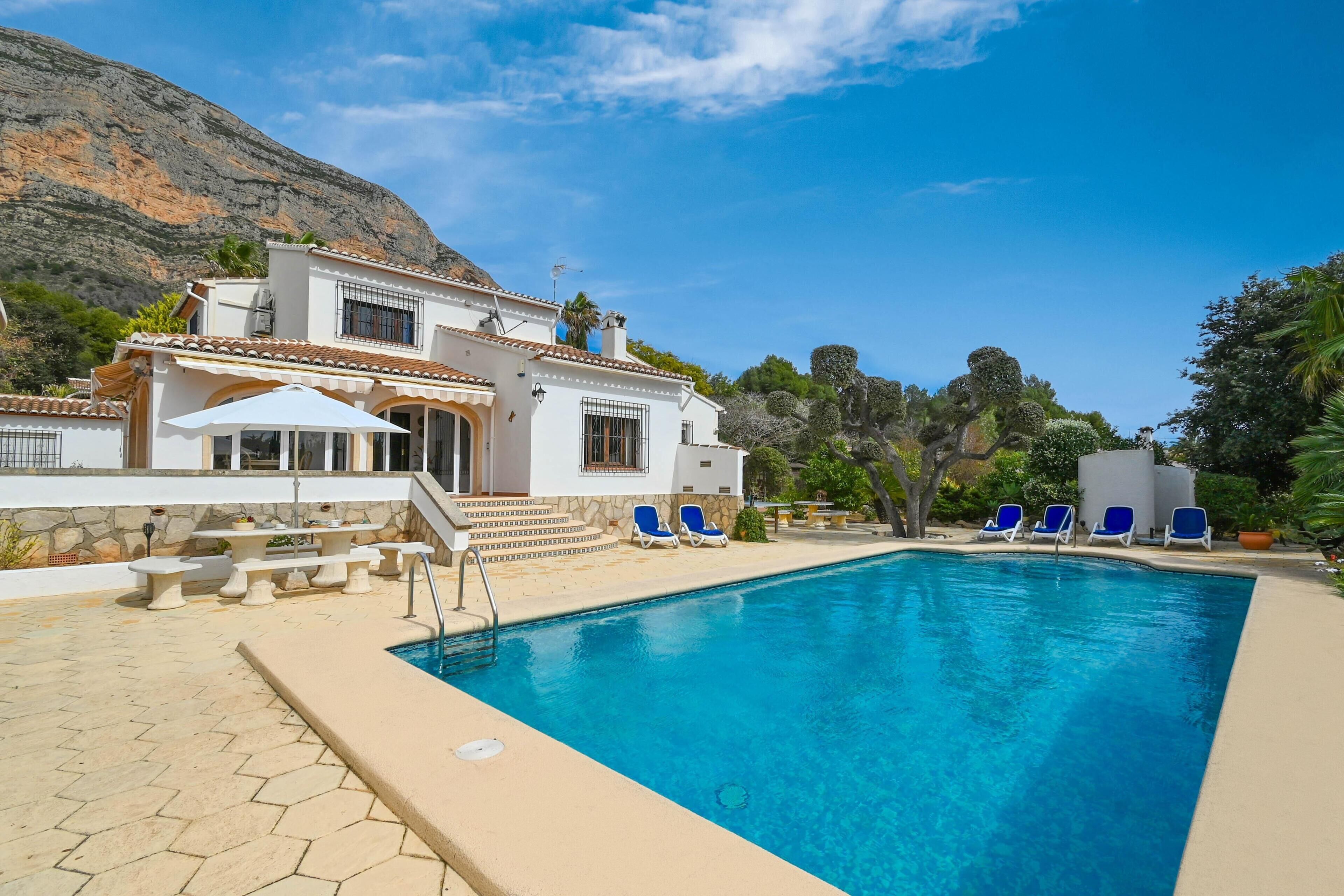 A pool area with a clear blue pool, white building with a tiled roof, and blue lounge chairs.