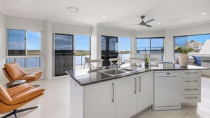 A modern kitchen with a central island, white cabinetry, and a dishwasher. There are two brown leather chairs and a dining table with chairs. Large windows offer views of the outdoors. - Admiralty Cove (Sunshine Coast)