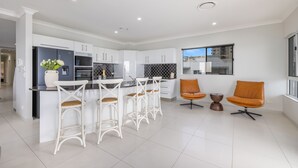A modern kitchen with white cabinets, a central island with bar stools, and a seating area with two orange chairs. - Admiralty Cove (Sunshine Coast)