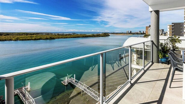 A balcony with a glass railing overlooking a calm body of water, a clear sky, and distant buildings. - Admiralty Cove (Sunshine Coast)