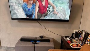 Un téléviseur fixé au mur, avec une télécommande et une console de jeux posée sur un meuble en dessous.
