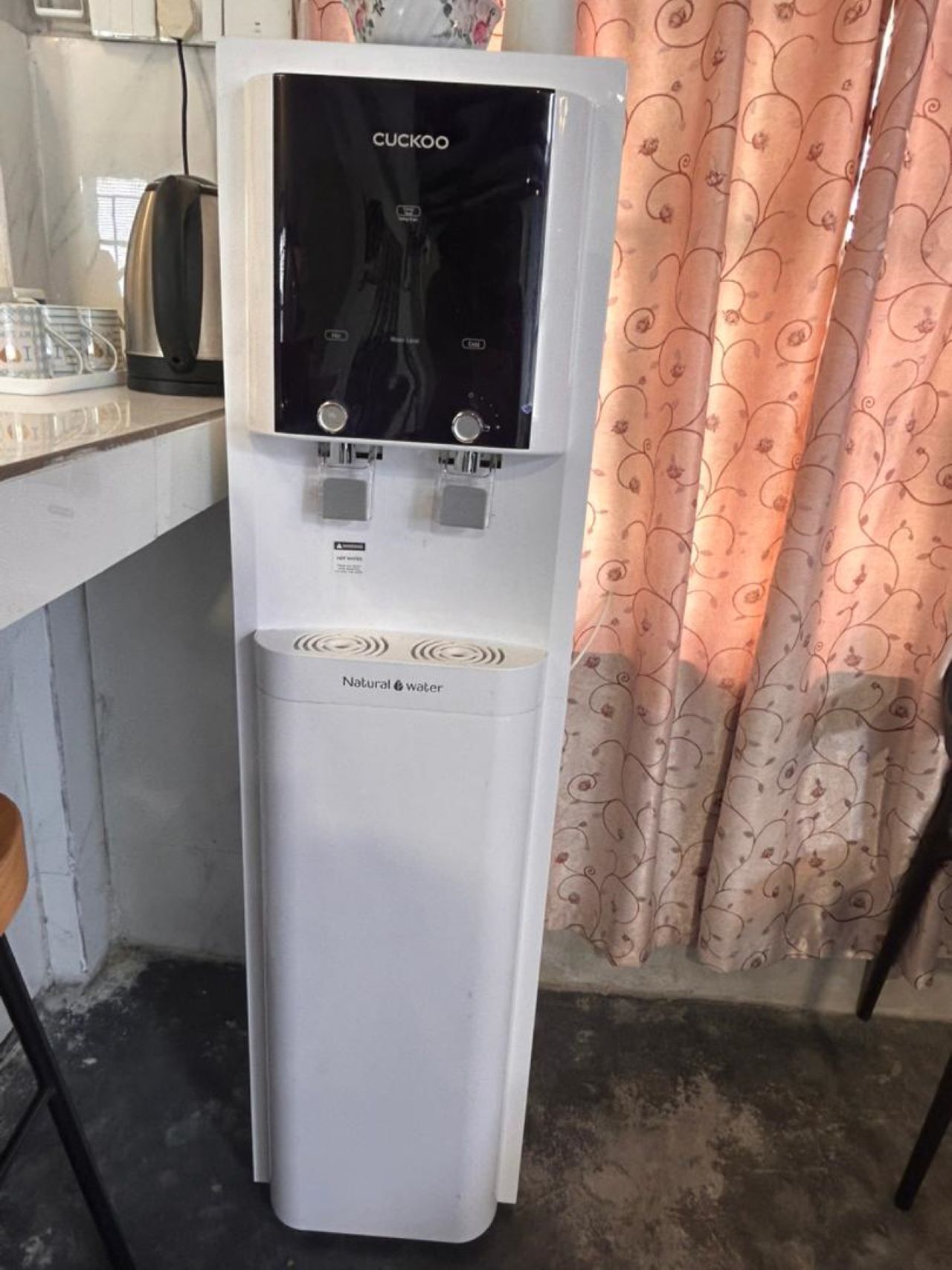 A Cuckoo water dispenser in a kitchen with a patterned curtain in the background.
