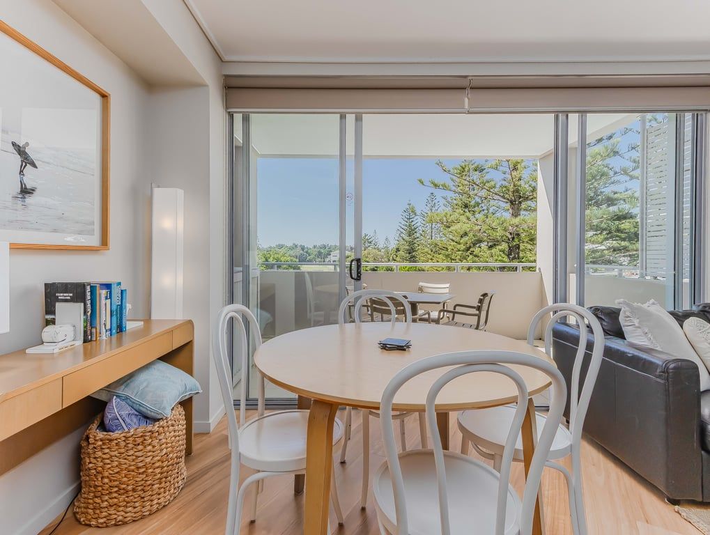 A modern dining area with a round table, white chairs, and a view of greenery through large windows.