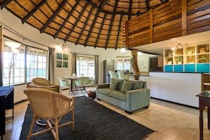 A spacious living area with a thatched ceiling, a sofa, wicker chairs, a dining table, and a kitchen area with a blue countertop. - Sable Ranch Selfsorg (Brits)