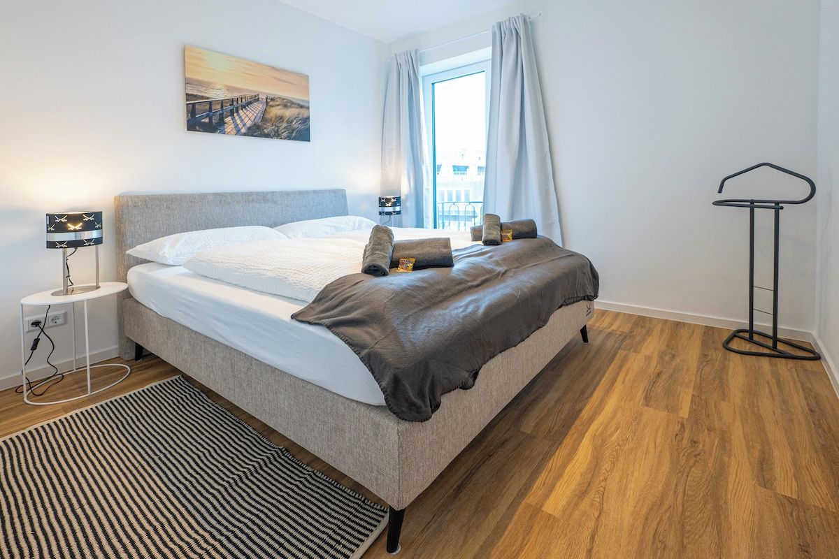 A bedroom with a bed, a bedside table, a lamp, a window with curtains, and a view of a building outside.