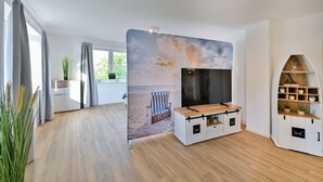 Ett modernt vardagsrum med en stor platt-TV, ett soffbord i trä, en bakgrund med strandtema och en inbyggd bokhylla.