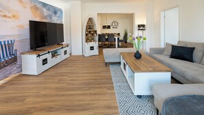 Ett modernt vardagsrum med en platt-TV, ett möblemang i trä för underhållningssystemet, en grå soffa och en matplats med en kristallkrona.