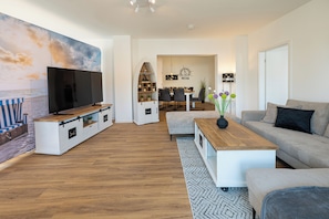 Ein modernes Wohnzimmer mit Flachbildfernseher, Holz-Entertainment-Möbel, grauem Sofa und einem Essbereich mit einer Deckenleuchte.
