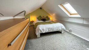 Quarto com teto inclinado, claraboia, cama com almofadas, mesa de cabeceira e mesa de apoio com abajur.