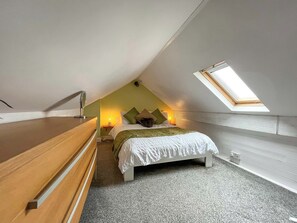 Een slaapkamer met een schuine plafond, dakraam, bed met kussens, nachtkastje en nachttafel met lamp.