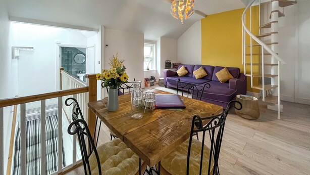 Uma sala de jantar com mesa de madeira, cadeiras, um vaso com flores e um sofá roxo com almofadas.