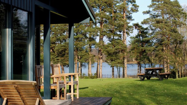 A covered outdoor seating area with wooden chairs and a picnic table, overlooking a lake with tall trees. - Conifer Chalet 5 - UKC5219 (Crieff)