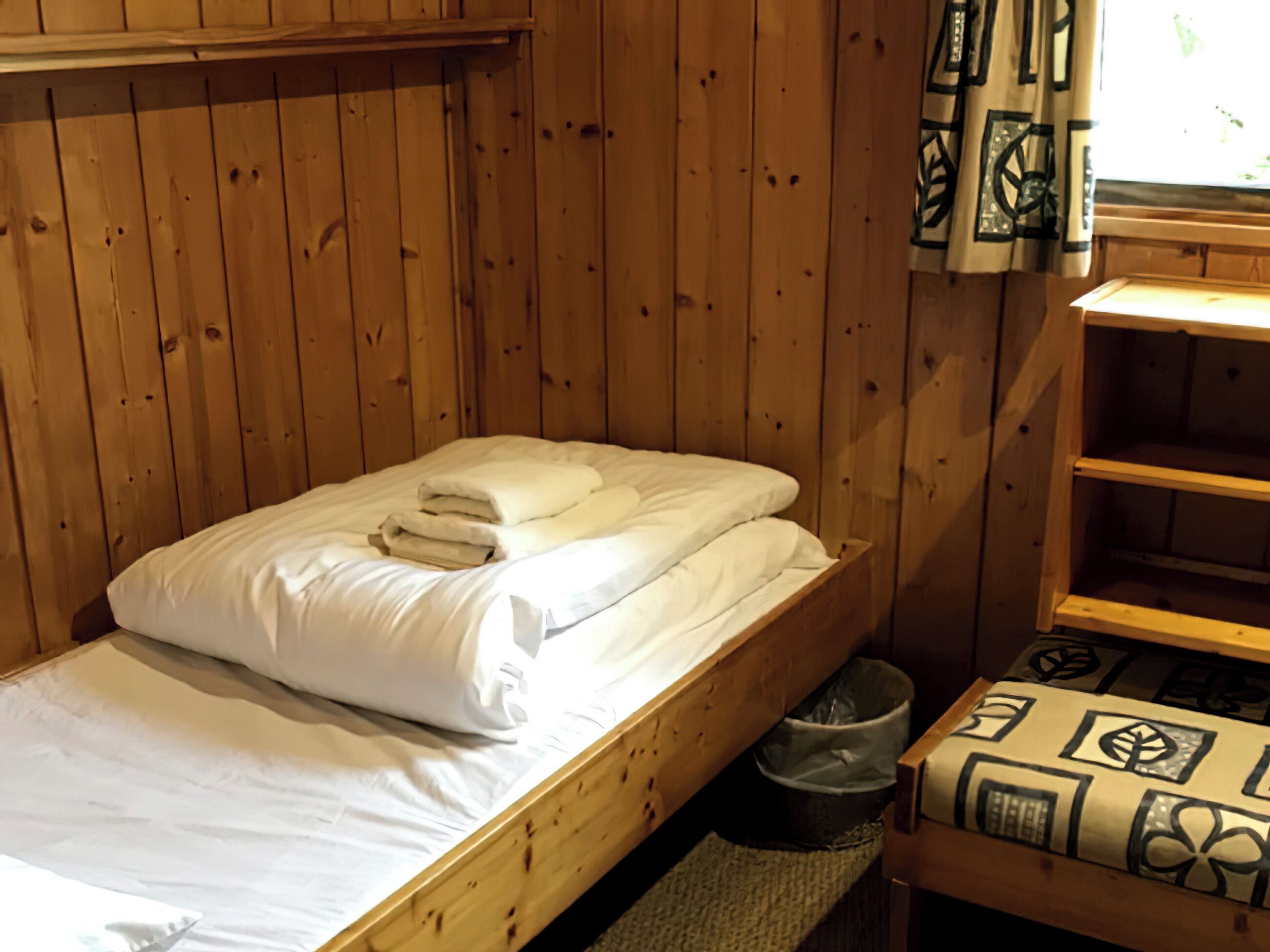 A single wooden bed with white bedding, a small trash bin, and a patterned cushion on a shelf.